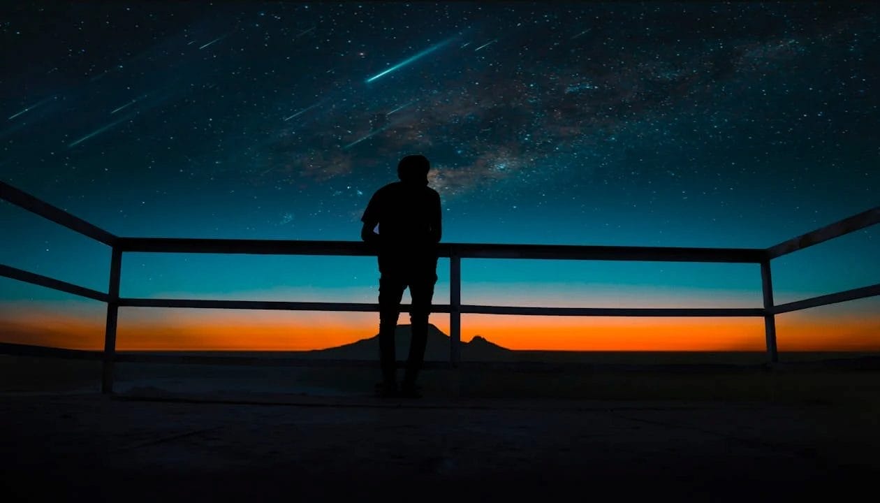 Silhouette of a person watching the Perseid meteor shower under a starry sky at twilight, with streaking meteors and a distant mountain view.