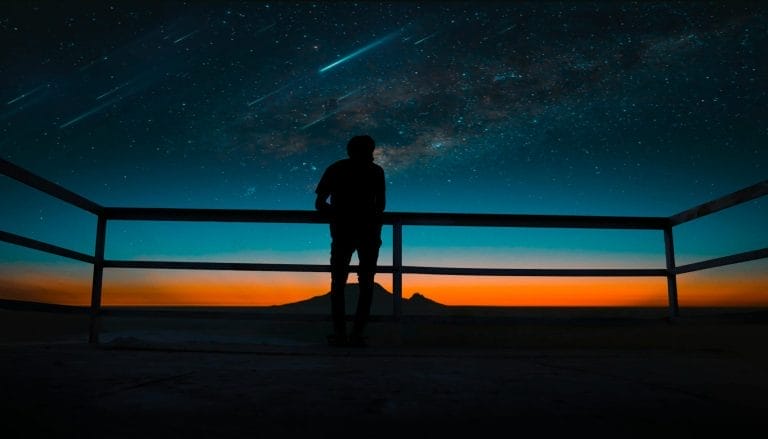 Silhouette of a person watching the Perseid meteor shower under a starry sky at twilight, with streaking meteors and a distant mountain view.